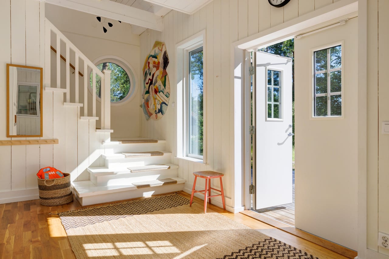 Vitmålad hall med trappa, jutemattor och öppen ytterdörr.