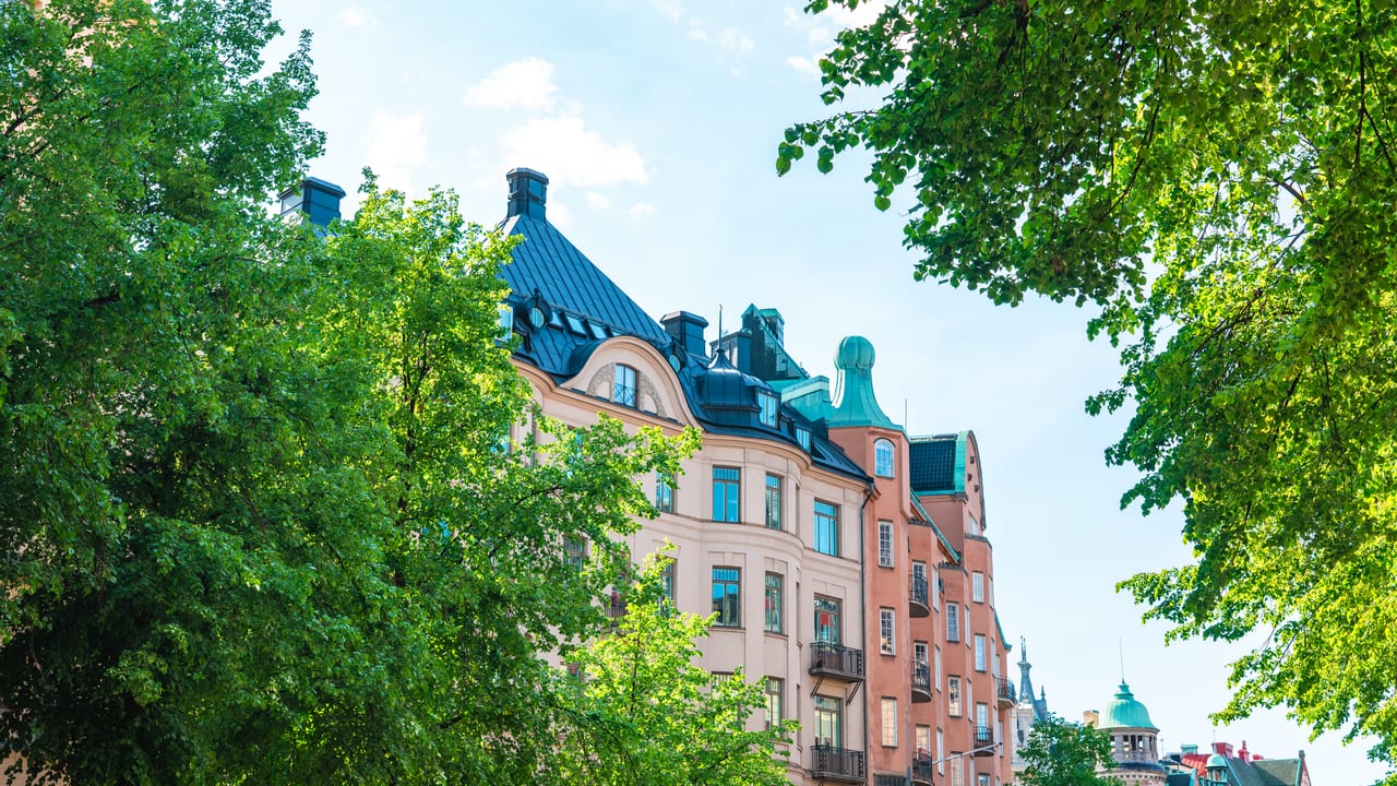 Putsade sekelskifteshus med kopparkupol bakom gröna lönnar.