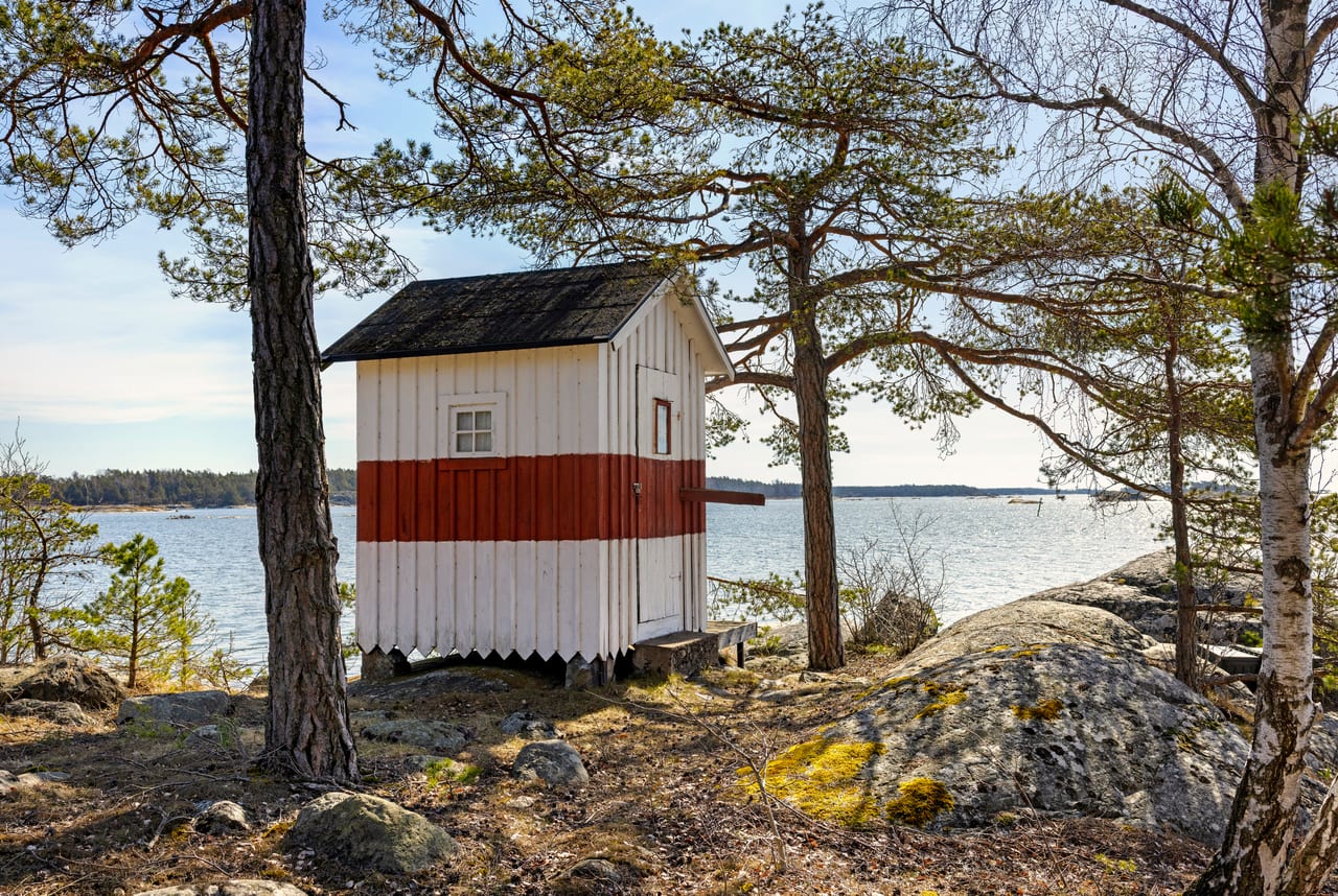 Röd och vit sjöbod mellan tallar vid en vik.